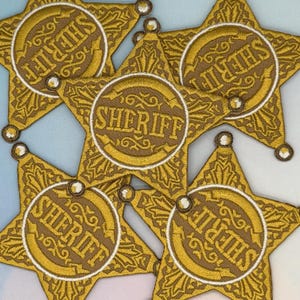 Op de afbeelding: Een verzameling geborduurde sheriff-badges. Elke badge heeft de vorm van een gouden ster met een cirkelvormig midden. Het woord "SHERIFF" is in het midden van elke badge geborduurd. De badges hebben een gedetailleerd, vintage ontwerp.