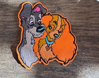 Puede incluir: Parche bordado con La Dama y el Vagabundo, una pareja de perros de dibujos animados. La Dama es un cocker spaniel marrón y blanco con un collar azul, y el Vagabundo es un perro callejero gris y blanco. El parche es naranja y tiene un contorno blanco.