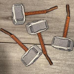 Może przedstawiać: Haftowane naszywki z młotem Thora, Mjolnirem. Każda naszywka przedstawia szarą głowicę młota z szczegółowym wzorem i brązowy uchwyt z pomarańczowymi akcentami w kształcie szewronów. Naszywki są ułożone na jasnej powierzchni z usłojeniem drewna.