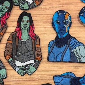 Puede incluir: Parches bordados con personajes de piel verde y pelo rosa, y piel azul. Los parches son de varios colores, incluyendo marrón, gris y azul. Los parches son probablemente para ropa o accesorios.