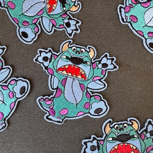 Könnte beinhalten: Gestickte Aufnäher mit einer Cartoon-Monsterfigur in Türkis, Blau, Lila und Rot. Die Aufnäher haben ein detailliertes Design mit sichtbaren Nähten und einem hellblauen Rand. Das Monster hat einen weit geöffneten Mund und befindet sich in einer dynamischen Pose.