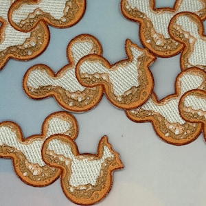 May include: Embroidered patches in a Mickey Mouse head shape. The patches have a white center with a textured appearance, bordered by a brown and orange outline. The patches are scattered across a light background.