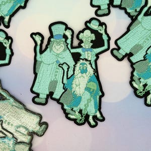 Puede incluir: Parches bordados que representan tres figuras fantasmales en tonos turquesa y azul, con bordes negros. Las figuras incluyen un fantasma con sombrero de copa, un fantasma esquelético y un fantasma barbudo, todos con costuras detalladas.