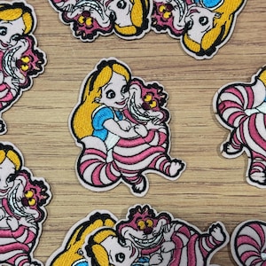 May include: Embroidered patches featuring Alice in Wonderland characters. The patches depict Alice in a blue dress and the Cheshire Cat in pink and white stripes. The patches have black borders and are set against a wood grain background.