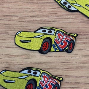 Puede incluir: Parche bordado amarillo con un coche de carreras de dibujos animados con el número 95 en rojo y azul. El coche tiene neumáticos negros con llantas rojas. Ideal para ropa o accesorios.