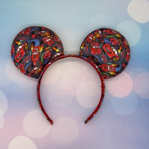 May include: A red headband with two large, round ears. The ears are patterned with images of the character Lightning McQueen from the movie Cars. The headband is red with a matching pattern on the ears.
