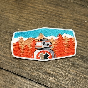 Könnte beinhalten: Ein gestickter Aufnäher mit BB-8, einer Figur aus Star Wars, der vor einem Wald mit blauem Himmel und weißen Bergen im Hintergrund steht. Der Aufnäher ist weiß, orange und schwarz.