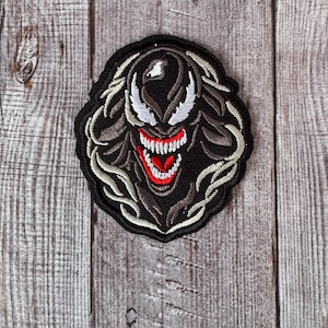 Op de afbeelding: Zwart-witte geborduurde patch van Venom, een personage uit Marvel Comics, met een rode mond en witte tanden. De patch is op een houten achtergrond.