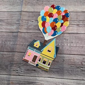 Può includere: Una casa gialla e rosa con un tetto nero è attaccata a un gruppo di palloncini colorati. La casa ha due finestre e una porta. I palloncini sono rossi, arancioni, gialli, verdi, blu e rosa.