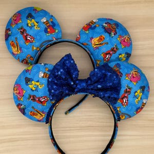 May include: A pair of mouse ear headbands with a blue sequined bow. The ears are blue with a repeating pattern of cartoon characters playing musical instruments. The headband is black with a matching pattern.