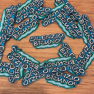 以下が含まれることがあります： 「SILENZIO BRUNO!」の文字が白で刺繍された、ティールとグリーンの背景のパッチのコレクション。パッチは木製の表面に散らばっています。