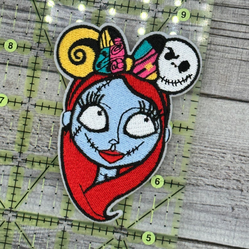 Puede incluir: Parche bordado con un personaje estilizado de piel azul, cabello rojo y detalles cosidos. El personaje tiene un lazo con acentos coloridos y una cabeza de Jack Skellington. El parche est&aacute; sobre un fondo de cuadr&iacute;cula.