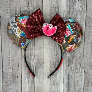 May include: Red and white sequined Minnie Mouse ears with a pink character from the film "Luca" on the bow. The ears are decorated with a colourful print of characters from the film.