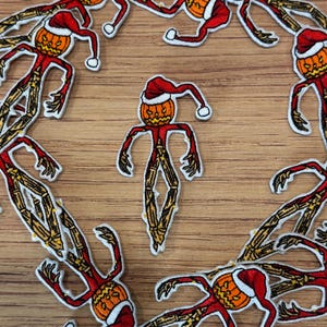 Könnte beinhalten: Gestickte Aufnäher mit einer vogelscheuchenartigen Figur mit einem Kürbiskopf, die einen roten Weihnachtsmannhut trägt. Die Figur hat rote Arme und einen Körper mit strohähnlichen Beinen. Die Aufnäher sind auf einer Holzoberfläche angeordnet.