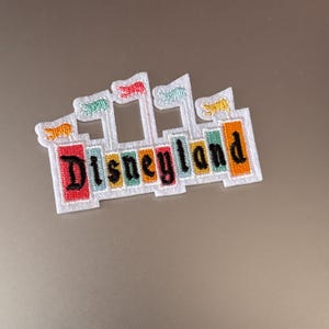 May include: A white embroidered patch with a colorful castle and the text "Disneyland" in black. The castle has five flags in various colors, including yellow, red, green, and blue.