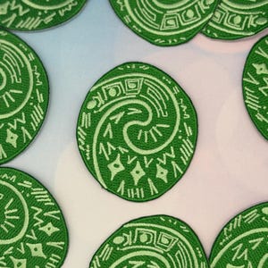 May include: Green embroidered patches with a white spiral and geometric design. The patches are circular and feature a variety of shapes and lines. The patches are likely for clothing or accessories.