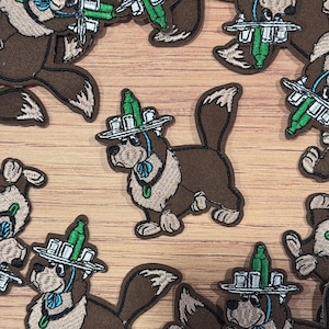 Może przedstawiać: Haftowane naszywki przedstawiające kreskówkowego psa w kapeluszu z zielonym wzorem butelki. Naszywki są brązowe, beżowe i zielone, z niebieskim kołnierzem. Naszywki są rozrzucone na jasnobrązowej drewnianej powierzchni.