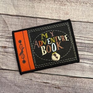 May include: A black and orange fabric patch with the text "My Adventure Book" in a colorful font. A small illustration of a jar with a cork is in the center of the patch.
