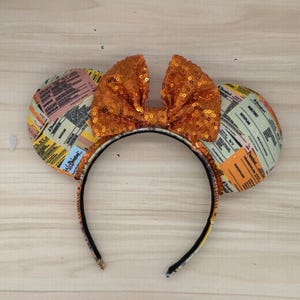 May include: A headband with mouse ears and a sequined bow. The ears are patterned with vintage-style tickets in shades of yellow, orange, and green. The bow is a vibrant orange color. The headband is black.