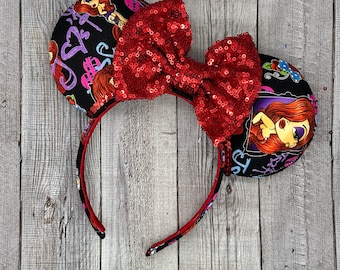 May include: A black headband with two round ears and a large red sequined bow. The ears are decorated with a cartoon illustration of a red-haired woman and colorful text on a black background. The headband is red.