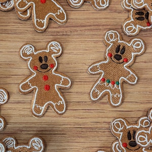 May include: Brown gingerbread bear patches with white trim, black eyes, and red accents. Some patches have red, green, and white details. The patches are scattered on a wooden surface.