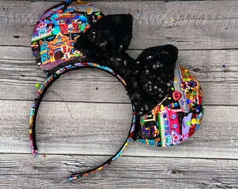 May include: A pair of black and multi-colored mouse ears with a black sequin bow. The ears are covered in a print of a theme park with various characters and attractions.