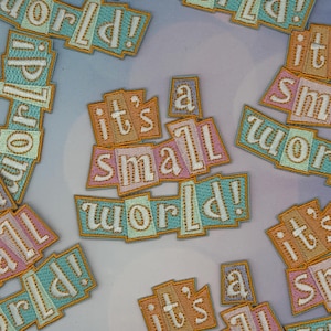 May include: Embroidered patches with the phrase "it's a small world!" in a colorful, whimsical font. The patches feature a variety of pastel colors, including orange, pink, and blue, with a gold border. The patches are scattered across a light background.