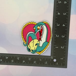 May include: Embroidered patch featuring a cartoon snake in teal, white, and red, holding a yellow skull. The snake is set against a pink heart-shaped background with a yellow scalloped border. The patch measures approximately 3 inches.