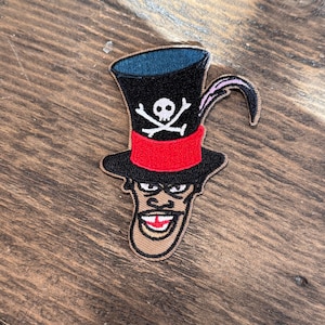 May include: Embroidered patch featuring a cartoon character wearing a black top hat with a red band and a skull and crossbones. The character has a tan face, a wide grin, and a pink feather. The patch is on a wooden surface.