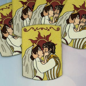 May include: Embroidered patches featuring a cartoon couple kissing, set against a yellow background with a sun design. The patches have a rectangular shape with a decorative border and are made with various colors.