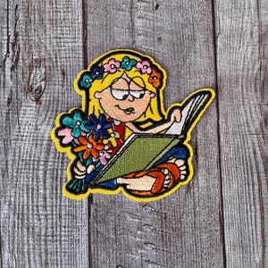 May include: A colorful embroidered patch of a girl with blonde hair wearing a flower crown and reading a book. The girl is wearing a yellow dress and has a bouquet of flowers in her hand.