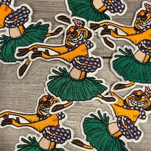 Op de afbeelding: Geborduurde patches met een stripfiguur in een hula rok en lei. Het personage is geel en bruin, met een brede grijns. De rok is groen en de lei is paars. De patches hebben een witte rand.