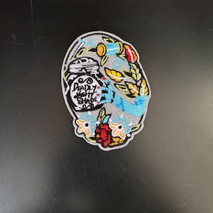 Könnte beinhalten: Ein grauer gestickter Aufnäher mit einem schwarzen Glas, auf dem "Deadly Night Shade" steht, und einer blauen Hand, die nach außen greift. Das Glas ist von bunten Blumen und Blättern umgeben.