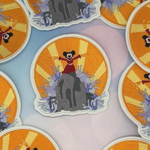 May include: A circular embroidered patch featuring a cartoon mouse in a red dress standing atop a gray rock formation. The background displays a bright orange sunburst pattern with light purple stylized waves crashing around the base of the rock.