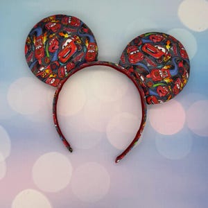 May include: Mickey Mouse ear headband featuring a Cars movie theme. The ears are patterned with images of Lightning McQueen and other characters on a gray background. The headband is red with a matching red interior.