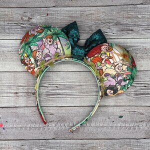 May include: A whimsical headband featuring two round ears with a colorful jungle scene print. The ears are adorned with a dark green sequined bow and a decorative vine. The headband is perfect for themed events or everyday wear.