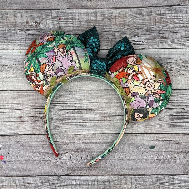 May include: A whimsical headband featuring two round ears with a colorful jungle scene print. The ears are adorned with a dark green sequined bow and a decorative vine. The headband is perfect for themed events or everyday wear.