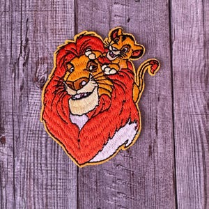May include: Embroidered patch featuring Simba and Mufasa from The Lion King. Simba is a small lion cub with a yellow mane, sitting on top of Mufasa, a larger lion with a red mane.