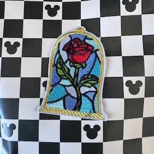 Puede incluir: Parche bordado con una rosa roja dentro de un diseño de vidriera. El parche tiene un borde dorado y está sobre un fondo de tonos azules, verdes y plateados. El parche está sobre una superficie a cuadros blancos y negros con siluetas de Mickey Mouse.