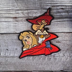 Pode incluir: Etiqueta bordada com uma figura com capuz vermelho com um pássaro azul, uma coruja marrom com bico branco e uma mulher loira com um vestido branco. A figura está usando uma capa vermelha e um chapéu vermelho com um esquilo marrom no topo.