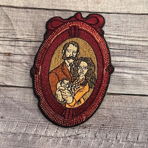 Może przedstawiać: Naszywka haftowana przedstawiająca portret rodzinny w ozdobnej, czerwonej ramie. Rodzina składa się z mężczyzny, kobiety i dziecka. Mężczyzna ma wąsy i jest ubrany w brązowy garnitur. Kobieta ma długie brązowe włosy i jest ubrana w żółtą sukienkę. Dziecko jest ubrane w biały śpioszek.