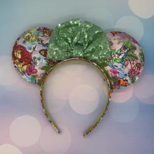 May include: A whimsical headband featuring mouse ears with colorful illustrations of animated characters and tropical flowers. The ears are connected by a green headband with a floral pattern. A large, sequined green bow sits atop the headband.