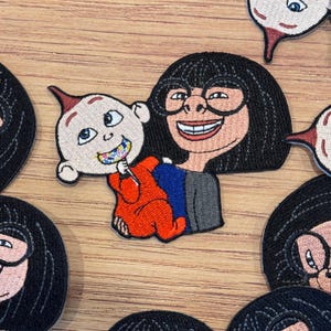 May include: Embroidered patch featuring Edna Mode and Jack-Jack from The Incredibles. Edna is smiling, wearing glasses, and has a black bob. Jack-Jack is in a red suit, with a colorful toothy grin. The patch is on a wooden surface.
