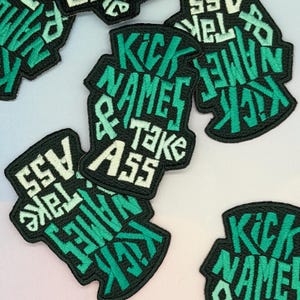 May include: Several black embroidered patches with the text "Kick Names & Take Ass" in a bold, stylized font. The text is mainly green with white highlights, set against a black backdrop. These patches are likely for clothing or accessories.