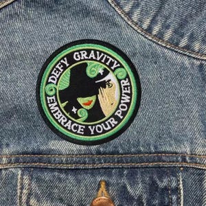 Puede incluir: Un parche redondo bordado con las palabras "Defy Gravity Embrace Your Power" en verde y blanco. El parche presenta una ilustración estilizada de una mujer con un sombrero negro y una cara verde. El parche está cosido a una chaqueta vaquera.