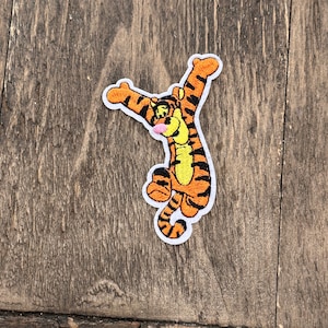 May include: An embroidered iron-on patch featuring a cartoon orange and black tiger with a pink nose, in a jumping pose with its arms raised.
