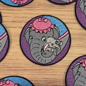 Könnte beinhalten: Ein gestickter Aufnäher mit einem grauen Elefanten mit einem rosa Hut und einem kleineren grauen Elefanten in seinem Rüssel. Die Elefanten sind von einem blauen und violetten Kreis umgeben.