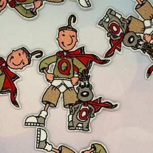 Op de afbeelding: Geborduurde patches met een stripfiguur in een superheldenpose, met een rode cape en een groen shirt met een grote "Q" erop. Het personage wordt vergezeld door een kleiner dier dat ook een cape draagt. De patches staan op een lichte achtergrond.
