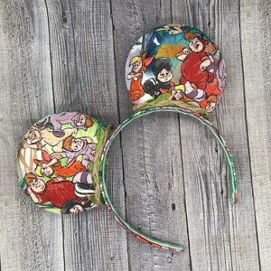 May include: A pair of decorative headband ears featuring a colorful Peter Pan theme. The ears are round with illustrations of characters from the movie. The headband is green with a floral pattern and the ears are attached with twine.