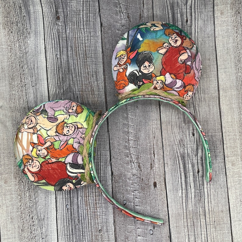May include: A pair of decorative headband ears featuring a colorful Peter Pan theme. The ears are round with illustrations of characters from the movie. The headband is green with a floral pattern and the ears are attached with twine.